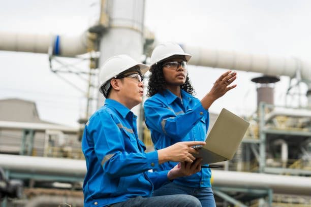 Engineers in hard hats and blue uniforms inspect industrial facility using a laptop.